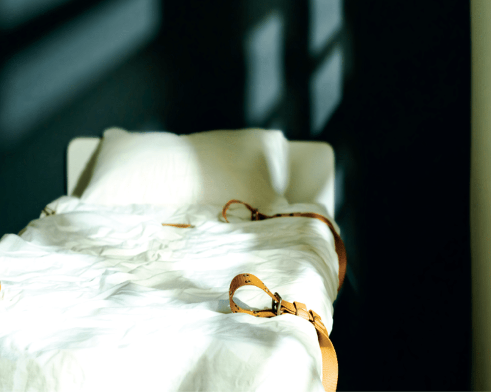 An empty white hospital bed with shackles next to a window. Shadows from the window frame cover the blank turquoise wall behind the bed.