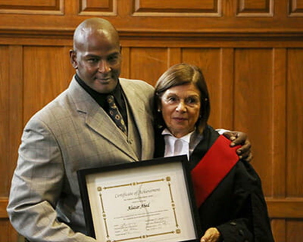 A man of colour in a suit and a Caucasian woman with short hair in a gown, both holding the graduation plaque smiling at the camera. 