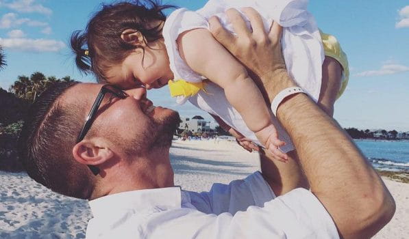 A man holding his baby daughter in the air making their foreheads touch at a beach.