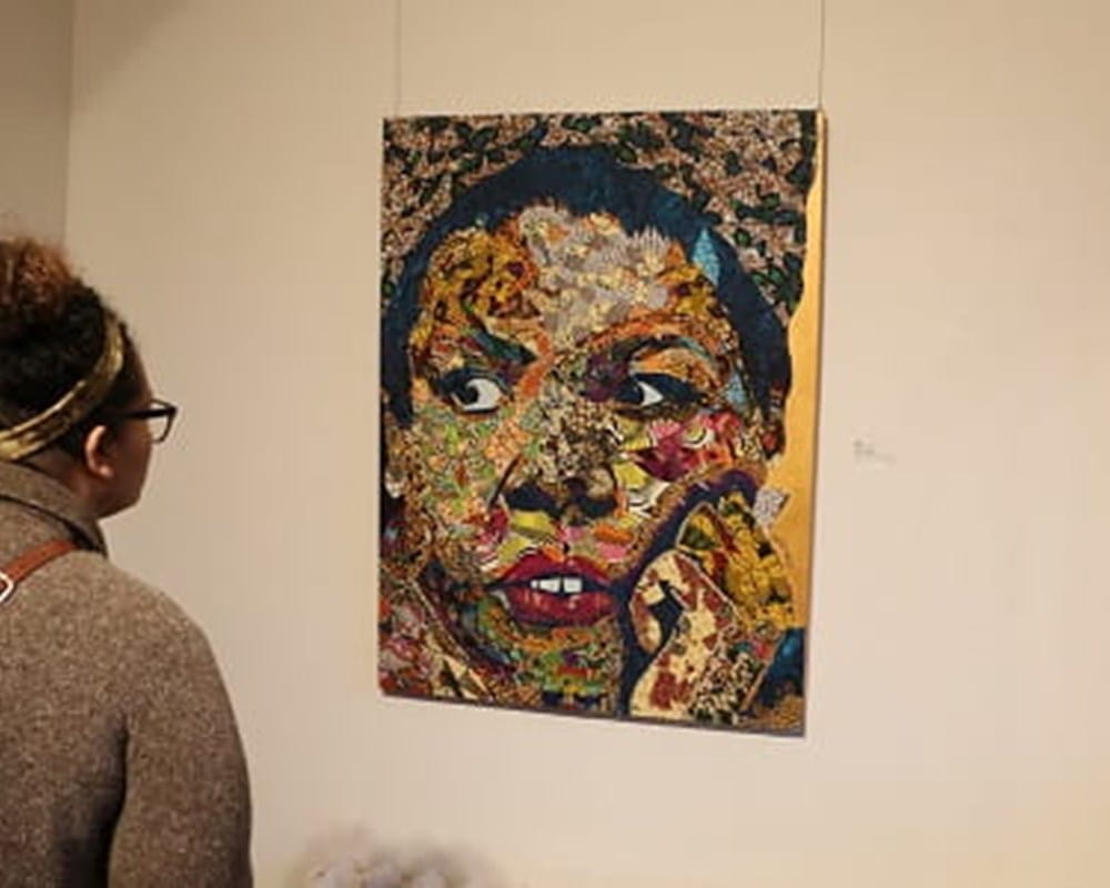 A woman of colour wearing a tan cardigan looking at an abstract piece of art hanging on a wall. 