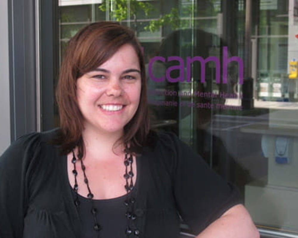 Cassandra wearing all black and a long black necklace posing beside the CAMH logo. 