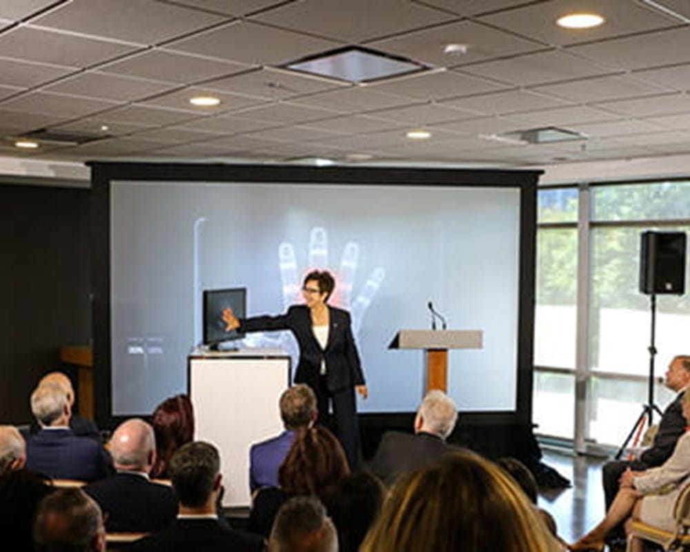 CEO, Catherine Zahn standing in front of an audience scanning her hand against a screen and it projecting it onto a screen. 