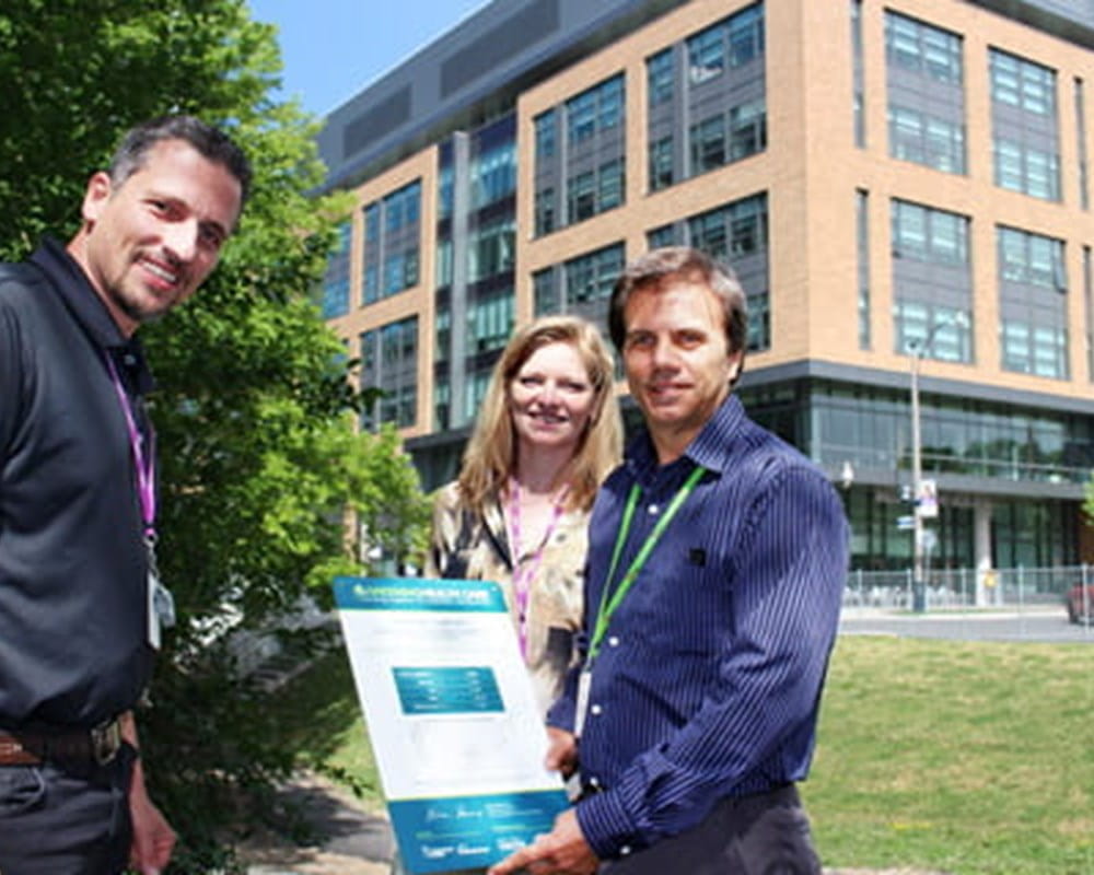Two CAMH staff standing in a field in front of the Bell Gateway Building with a man in a green lanyard holding a blue and white plaque. 