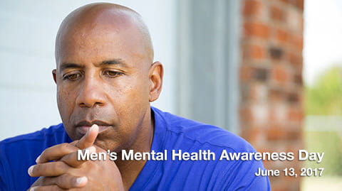 A bald man of colour wearing a blue t-shirt clasping his hands in front of his face looking upset. 