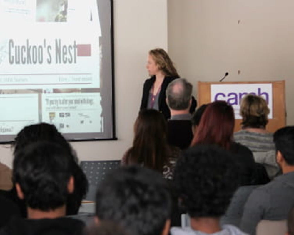 A woman presenting in front of a large diverse crowd with a projected image of a newspaper headline 