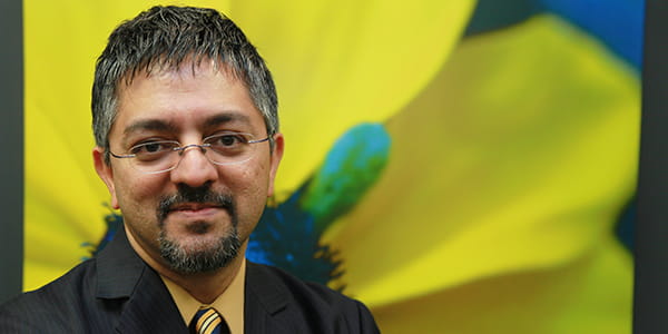 A coloured man with glasses smiling in front of an image of a flower with yellow petals. 