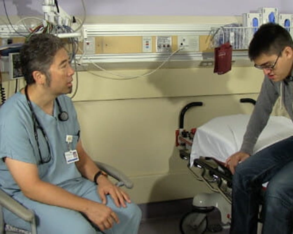 A patient sitting on a hospital bed and a doctor in green scrubs talking to him. 