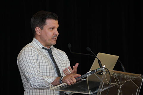 A man in a white and grey checkered button shirt and black tie speaking in front of a clear podium. 