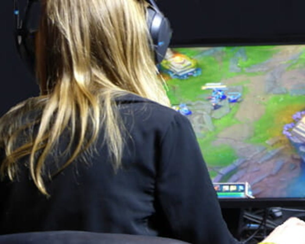 A person with medium length dirty blond hair wearing headphones sitting in front computer screen playing a video game. 