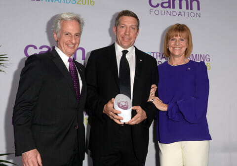 Two men in business suits and a woman in a blue blouse standing beside each other smiling while holding a white award. 