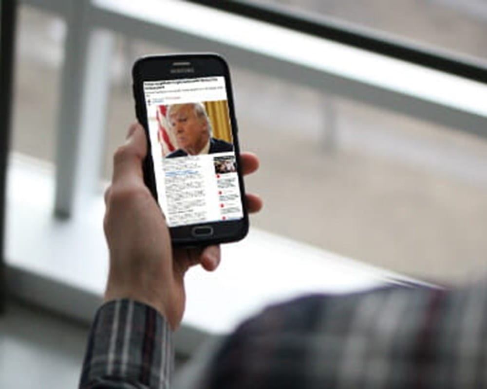 A man wearing a long sleeve plaid shirt holding a phone with a picture of Donald Trump in a news headline. 