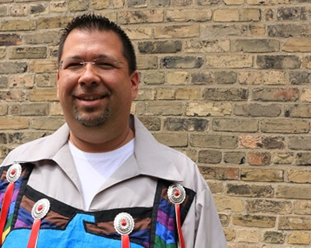 A man of Aboriginal descent smiling in front of a brick wall. 