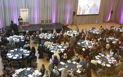 Several black round tables with people dressed in business clothes sitting around them, watching a person speaking in front of the podium on stage. 